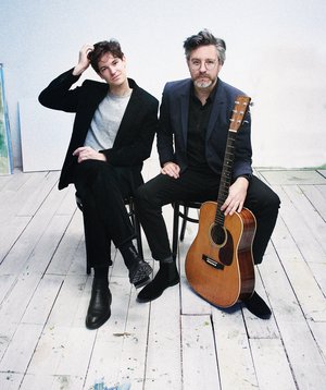 Die beiden Musiker sitzen vor einer weißen Wand auf Stühlen.