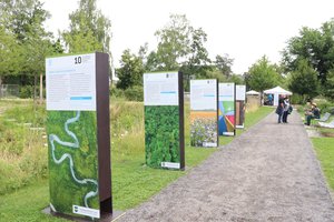 Ausstellungstafeln zum thema Klimaschutz, die neben einem Weg stehen. Daneben eine Grünanlage, im Hintergrund mehrere Menschen