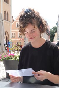Junge Frau liest im Freien eine Karte, im Hintergrund ein Blumentopf und Häuser der Stadt