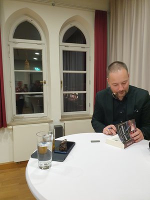Ein Mann, der Autor Tom Hillenbrand, steht im Foyer vor dem Festsaal des Rahtauses an einem Stehtisch und signiert sein Buch.